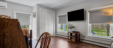 Una habitación con suelo de madera, una silla de madera, una puerta blanca, un televisor montado en la pared y una ventana con vistas a un paisaje verde.
