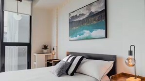 Une chambre d’hôtel moderne, équipée d’un lit, d’une table de chevet avec une lampe, d’un tableau au mur et d’une fenêtre avec des stores.