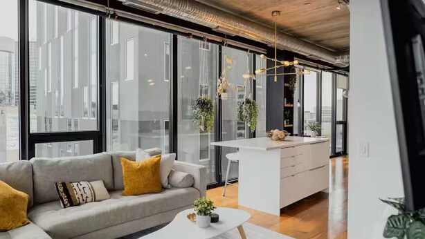 Un espace de vie moderne comprenant un canapé, une table basse et un coin cuisine doté d’un plan de travail blanc et de plantes suspendues.