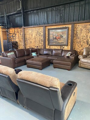 Un vaste espace de vie comprenant un canapé d’angle, des poufs, un fauteuil en cuir et un mur décoratif en bois agrémenté d’un tableau encadré.