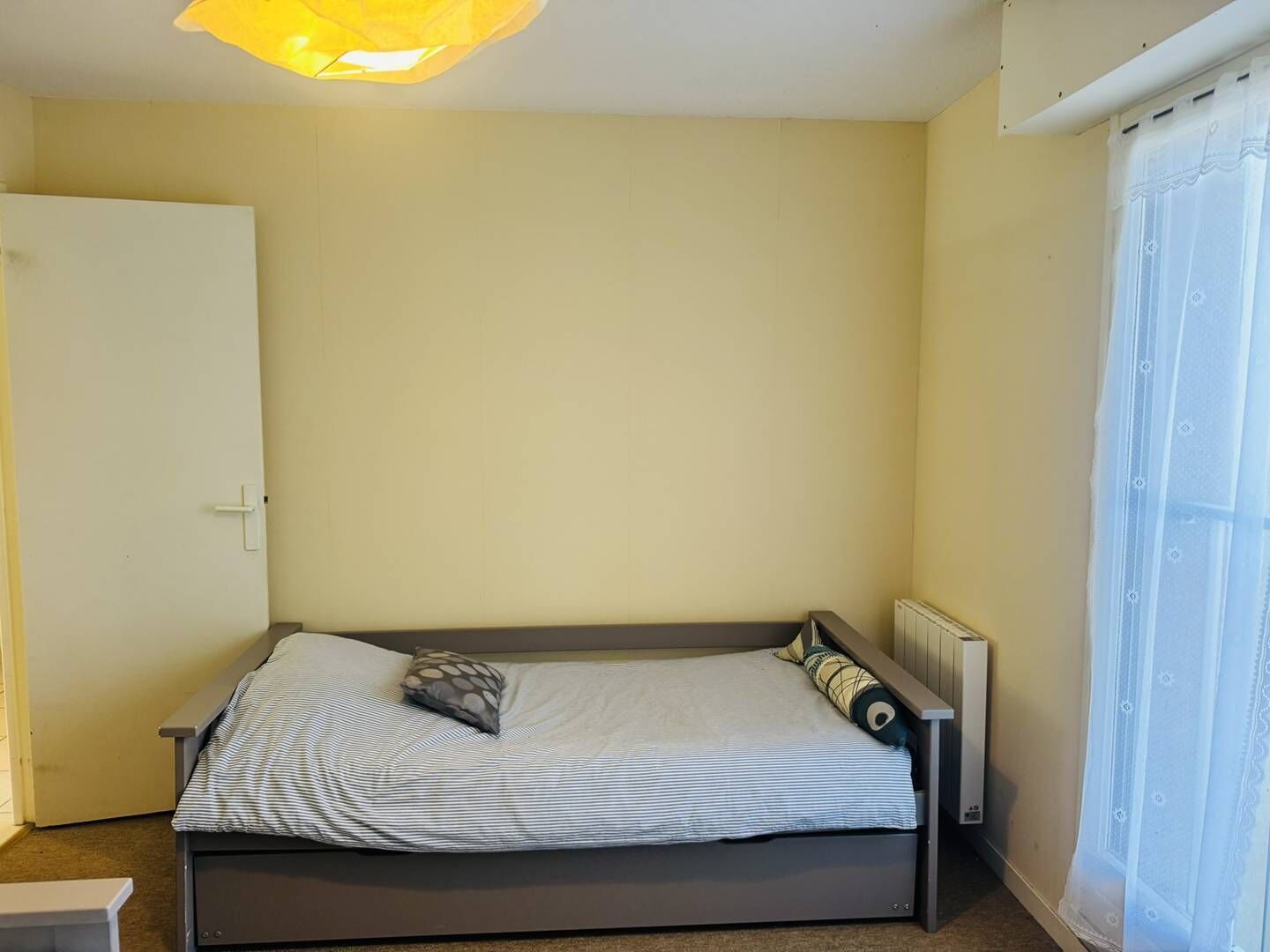 A single bed with a striped bedspread and two pillows, a white door, a radiator, and a window with sheer curtains.