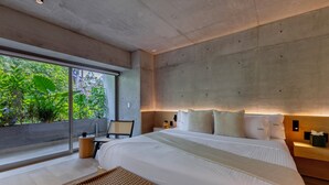 Ein modernes Schlafzimmer mit einem großen Bett, einem Nachttisch, einem Sessel und Blick ins Grüne durch ein großes Fenster.