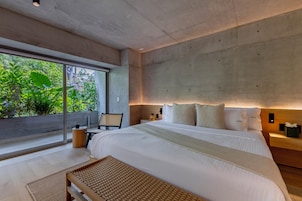 A modern bedroom with a large bed, a bedside table, a chair, and a view of greenery through a large window.