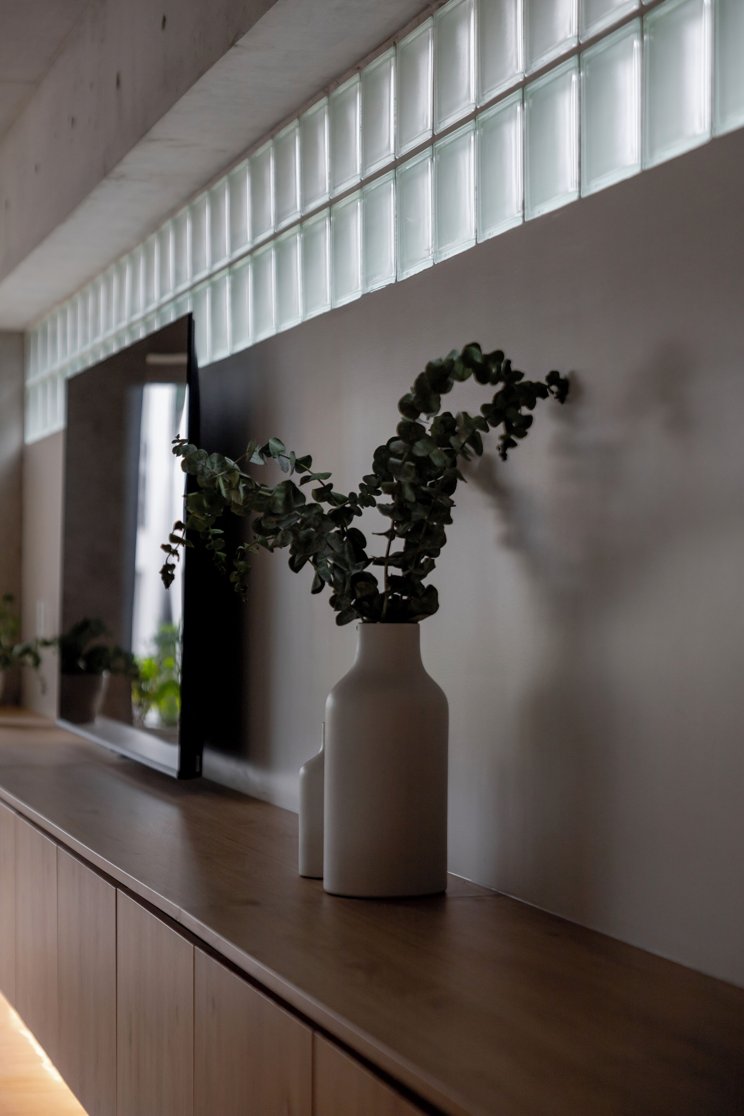 A white vase with a plant on a wooden counter against a wall with a window.
