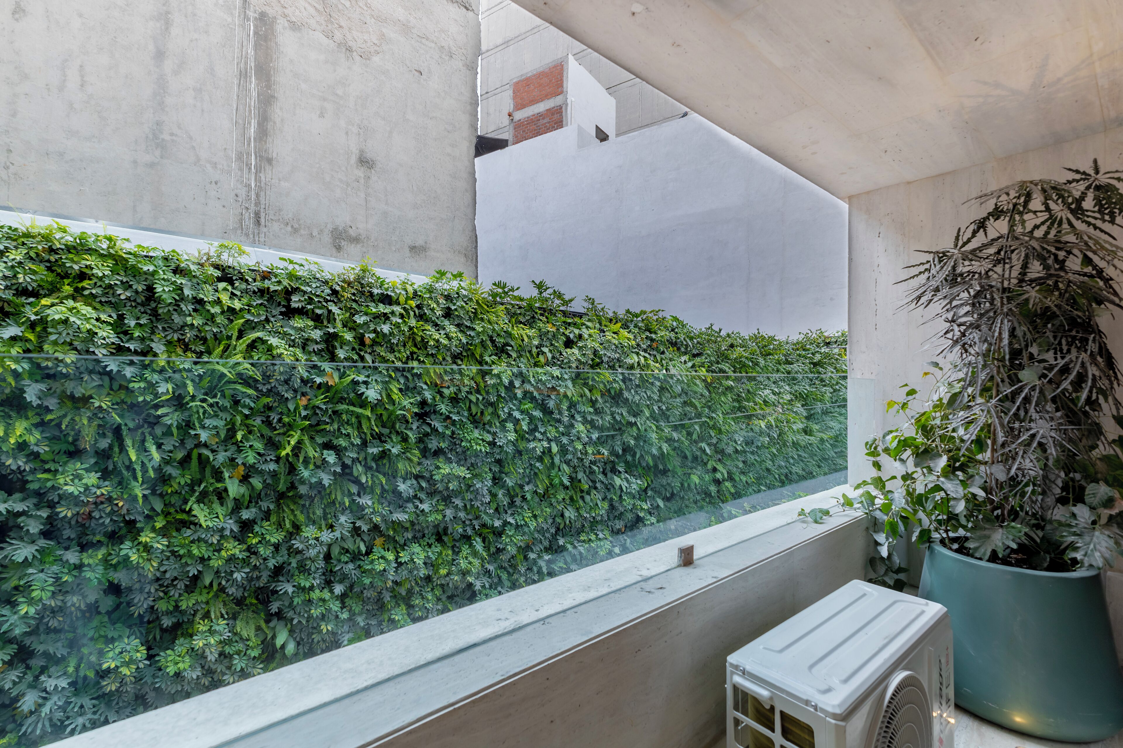 Un balcón con un seto verde, una planta en maceta y un equipo de aire acondicionado blanco.