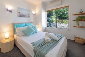 A neatly made bed with a white and teal color scheme, a bedside lamp, a window with blinds, and a wall-mounted shelf with decorative items.