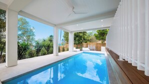 Une piscine dotée d’une terrasse en bois, de colonnes blanches et offrant une vue sur les arbres et une clôture.