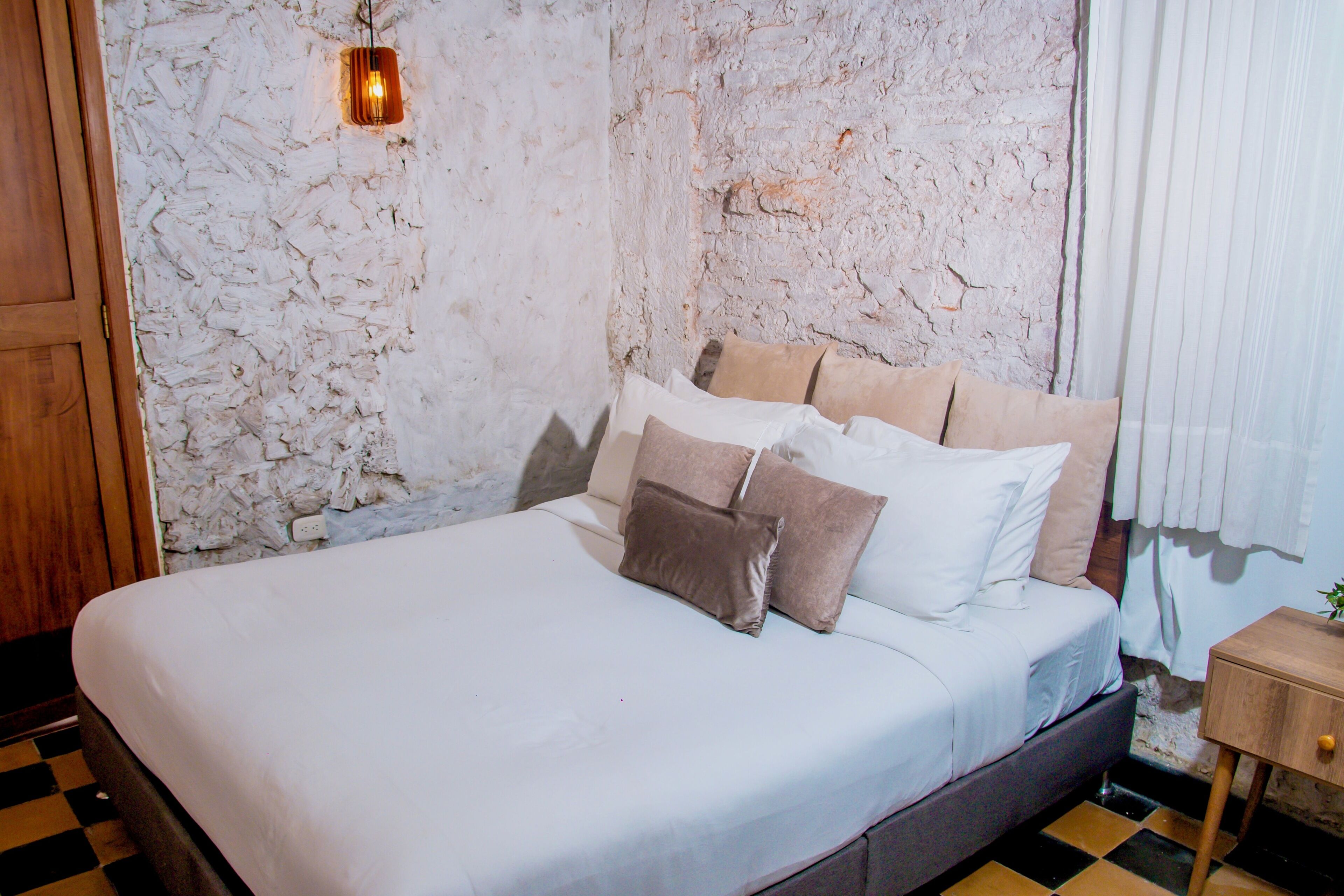 A bed with white bedding and multiple pillows, a wooden nightstand with a plant, and a textured wall.