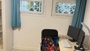 Ein Zimmer mit einem Bett, einem Schreibtisch mit Computer, einem Stuhl mit Blumenkissen und einem Fenster mit Vorhängen.