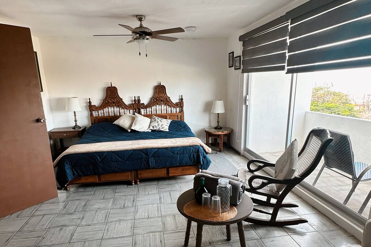 Un dormitorio con cama, ventilador de techo, ventana con persianas, una mecedora y una pequeña mesa con objetos encima.