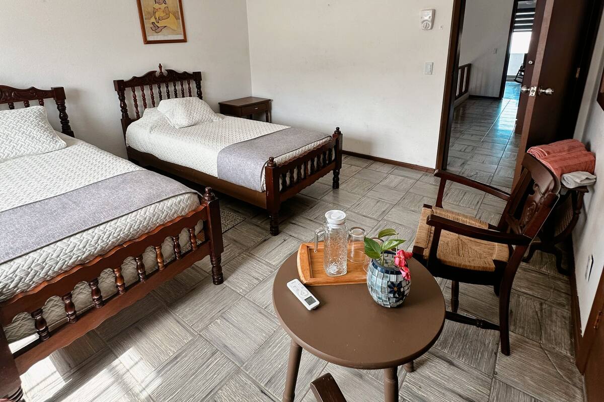 Una habitación con dos camas de madera, un ventilador de techo, una mesita con un teléfono, una silla y una puerta que da a otra habitación.