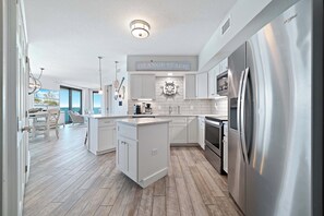A modern kitchen with white cabinetry, a stainless steel refrigerator, and a sign reading 'Orange Beach.' - Summer House 1101B | Beachfront Condo | Resort Amenities | Indoor & Outdoor Pool | My Beach Getaways (Orange Beach)