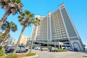 A multi-story hotel with a modern design, featuring a large parking area with several vehicles, palm trees, and a clear blue sky. - Seawind 1804 | Beachfront | Pool | Hot Tub | My Beach Getaways (Gulf Shores)