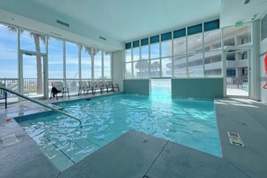 An indoor swimming pool with clear glass walls, a poolside area with chairs, and a view of the outside. - Seawind 1804 | Beachfront | Pool | Hot Tub | My Beach Getaways (Gulf Shores)