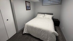 Een slaapkamer met een bed, nachtkastje, deur, foto aan de muur en een raam met jaloezieën.