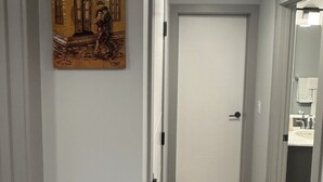 Een hal met een witte deur, een plafondlamp en een kunstwerk aan de muur dat een stadsgezicht toont.