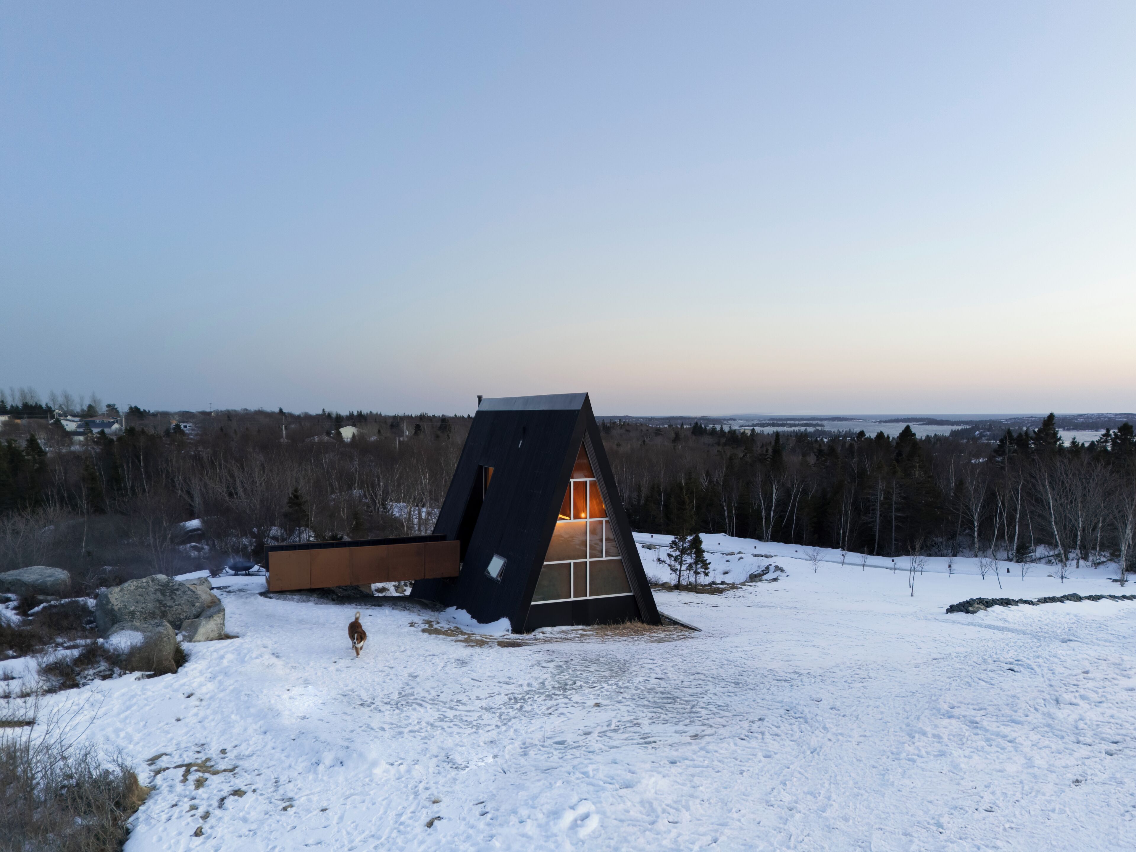 A modern cabin with a sloped roof, surrounded by a snowy landscape and a view of distant trees.