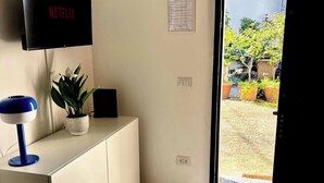 Ein Zimmer mit Fernseher an der Wand, einem Schreibtisch mit Lampe, einem Blumentopf und einer Tür zum Außenbereich.