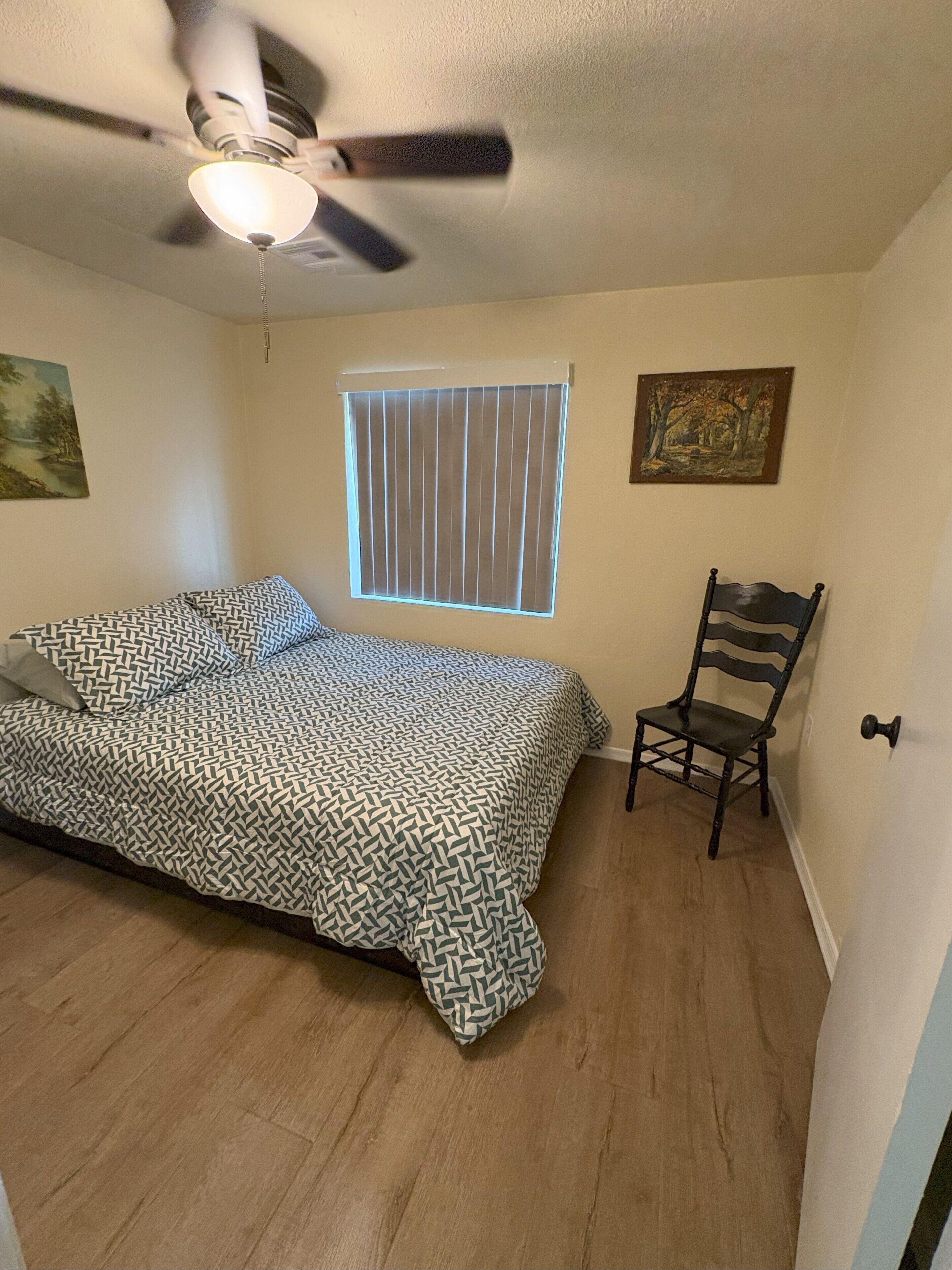 A bedroom with a bed, a chair, a window with blinds, a ceiling fan, and two wall-mounted paintings.