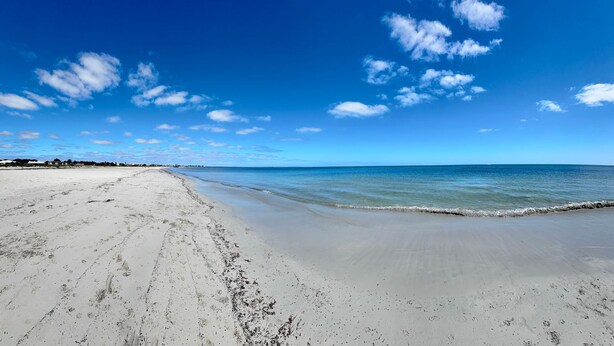 Une vaste plage de sable fin, avec de douces vagues, un ciel bleu clair et quelques nuages épars.