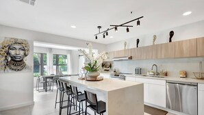 A modern kitchen with a central island, bar stools, a dining area, and a view through large windows. - Modern Home • Pool • Near Beach & Las Olas (Fort Lauderdale)