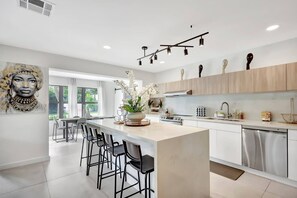 A modern kitchen with a central island, bar stools, a dining area, and a view through large windows. - Modern Home • Pool • Near Beach & Las Olas (Fort Lauderdale)