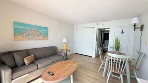 Ein Wohnzimmer mit Sofa, Couchtisch und Esstisch mit Stühlen. An der Wand hängt ein Gemälde, es gibt eine Lampe und eine Tür zu einem anderen Raum.
