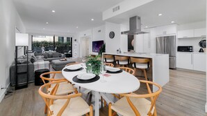 Un salon moderne avec une table à manger pour quatre personnes, une cuisine dotée de meubles blancs et d’appareils électroménagers en acier inoxydable, et une grande fenêtre offrant une vue sur la verdure.