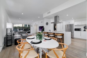 A modern living room with a dining table set for four, a kitchen with white cabinets and stainless steel appliances, and a large window offering a view of greenery. - Hollywood Hills, Runyon Canyon Townhouse (Los Angeles)