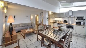 A modern kitchen with built-in appliances, a dining area with a wooden table and chairs, and a living room with a sofa and a lamp. - PAPRIKA - Villa Port Nature 69 - Luxury - 4 people (AGDE)