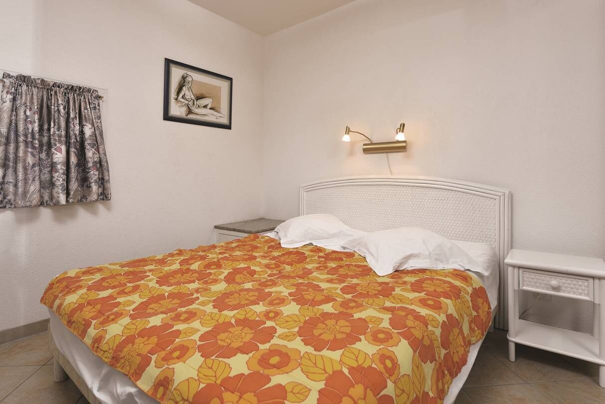 A bedroom with a bed, a nightstand, a wall-mounted light, a framed picture, and a window with curtains.