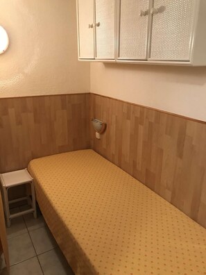 Una cama individual con una colcha estampada, un taburete pequeño y un inodoro fijado a la pared.