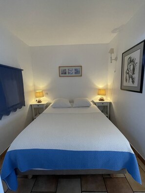 Een eenpersoonsbed met een wit-blauwe sprei, twee bedlampjes, een ingelijst schilderij boven het bed en een wandlamp aan de rechterkant.