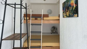 Ein kleines Zimmer mit Etagenbetten, einer Metallleiter, einem kleinen Ventilator und einem farbenfrohen Bild an der Wand.