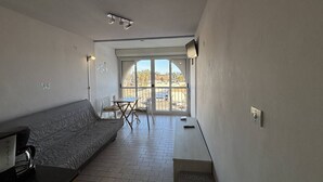 Ett kompakt vardagsrum med en soffa, ett litet bord, en stol och en skjutdörr i glas som leder ut till ett utomhusområde.