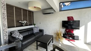 A modern living room with a black leather sofa, a black coffee table, a mounted flat-screen TV, and a large window. - VILLA SAVANNAH - Villa Port Nature 105 - Luxury - 4 people (AGDE)