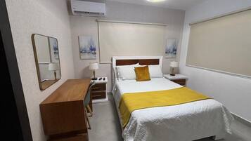 Une chambre d’hôtel équipée d’un lit, d’un bureau, d’une chaise, d’une table de chevet, d’une lampe et d’un climatiseur fixé au mur.