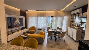 A modern living room with a sofa, dining table, and chairs, a flat-screen TV, and a kitchen area with a stove and cabinets. - De Lujo (Santiago de los Caballeros)