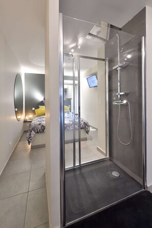 Une salle de bain moderne avec une douche à parois de verre, des toilettes apparentes et un lit avec une literie à motifs dans la pièce adjacente.