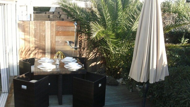 Um pátio externo com uma mesa posta para quatro pessoas, rodeado por cadeiras de vime, uma cerca de madeira e uma palmeira.
