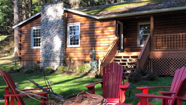 A log cabin with a stone chimney, a porch with red Adirondack chairs, and a fire pit.