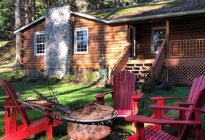 Une cabane en rondins avec une cheminée en pierre, une véranda avec des fauteuils Adirondack rouges et un foyer extérieur.