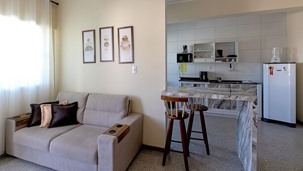 En stue med sofa, sofabord og et køkkenområde med køleskab, mikroovn og barstole.