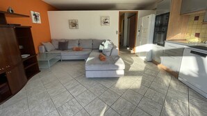 Een woonkamer met een bank, een voetenbankje en een houten kast. Er is een keuken met een koelkast en fornuis.