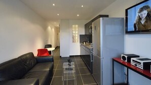 Una sala de estar moderna con un sofá de cuero negro, una mesa de centro de vidrio transparente, un área de cocina con un refrigerador y un televisor de pantalla plana montado en la pared.
