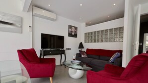 Een moderne woonkamer met een flatscreen televisie, rode fauteuils, een glazen salontafel en een grijze bank met decoratieve kussens.