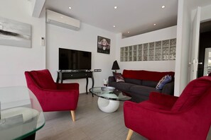 Ein modernes Wohnzimmer mit Fernseher, roten Sitzmöbeln, einem Glastisch und einem grauen Sofa mit Zierkissen.