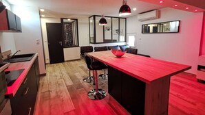 A modern kitchen with a wooden island, black bar stools, and a red accent wall. - VOLUPTIA  - Villa Port Nature 54 - Luxury - 4 people (AGDE)