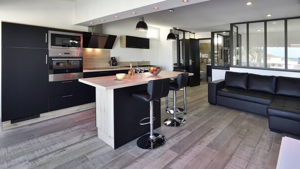 A modern kitchen with a central island, black cabinetry, stainless steel appliances, and a black leather sofa in the living area. - VOLUPTIA  - Villa Port Nature 54 - Luxury - 4 people (AGDE)
