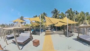 Une zone aménagée au bord de la plage, avec plusieurs espaces de repos ombragés, dont certains sont dotés de bancs et d’autres de tables, le tout entouré de palmiers.